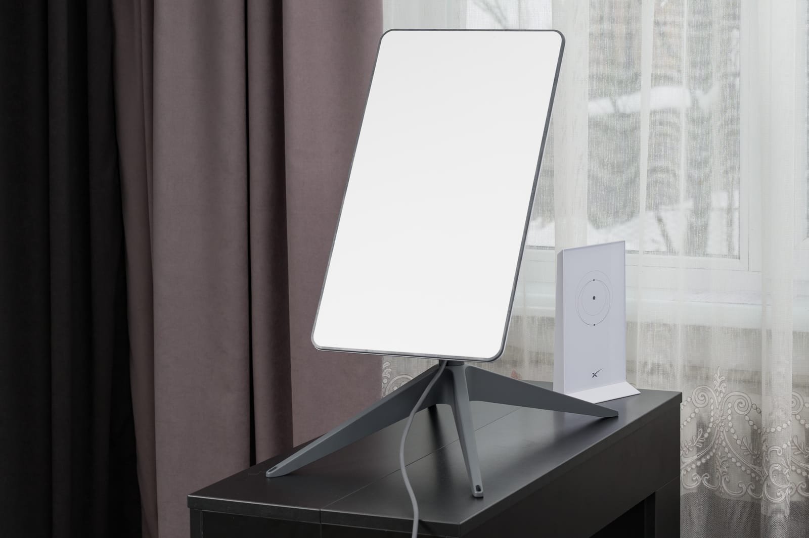 Pravokutna satelitska antena Starlink Hrvatska na stalku stoji na crnom stolu pored bijelog elektroničkog uređaja, ispred prozora s prozirnim zavjesama, naglašavajući dostupnost i modernu tehnologiju.