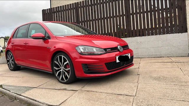 Crveni Volkswagen Golf GTI hatchback parkiran je na asfaltiranom prilazu pored drvene ograde i bijelog zida, svježe održavan nakon velikog servisa u sklopu redovnog servisnog rasporeda Golfa 7.