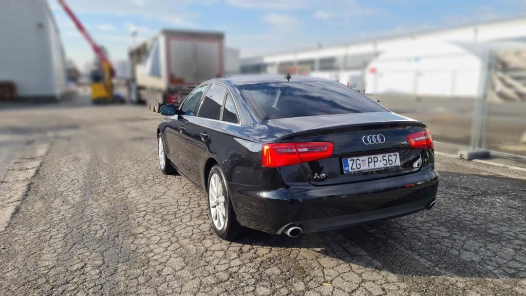 Crna Audi A6 limuzina s PP tablicama parkirana je na ispucaloj asfaltnoj površini u blizini industrijskih zgrada pod djelomično oblačnim nebom.