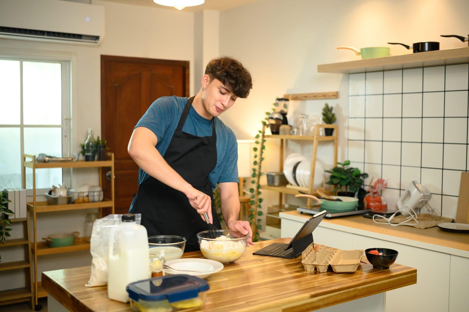Osoba u pregači miješa smjesu za palačinke u zdjeli u modernoj kuhinji, pažljivo slijedeći recept za palačinke prikazan na ploči na pultu.