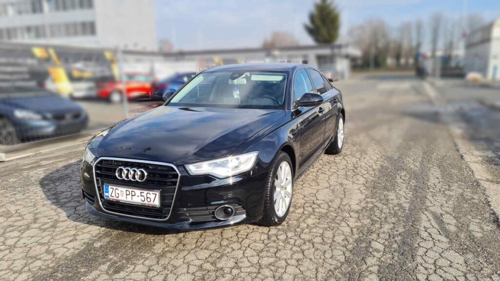 Crni Audijev sedan s PP pločama parkiran je na ispucalom pločniku na vanjskom parkiralištu, sa zgradama i ogradom u pozadini.