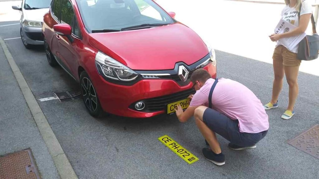 Muškarac kleči kako bi pričvrstio žutu registarsku pločicu na prednji dio crvenog Renaultovog automobila dok žena stoji u blizini na ulici.