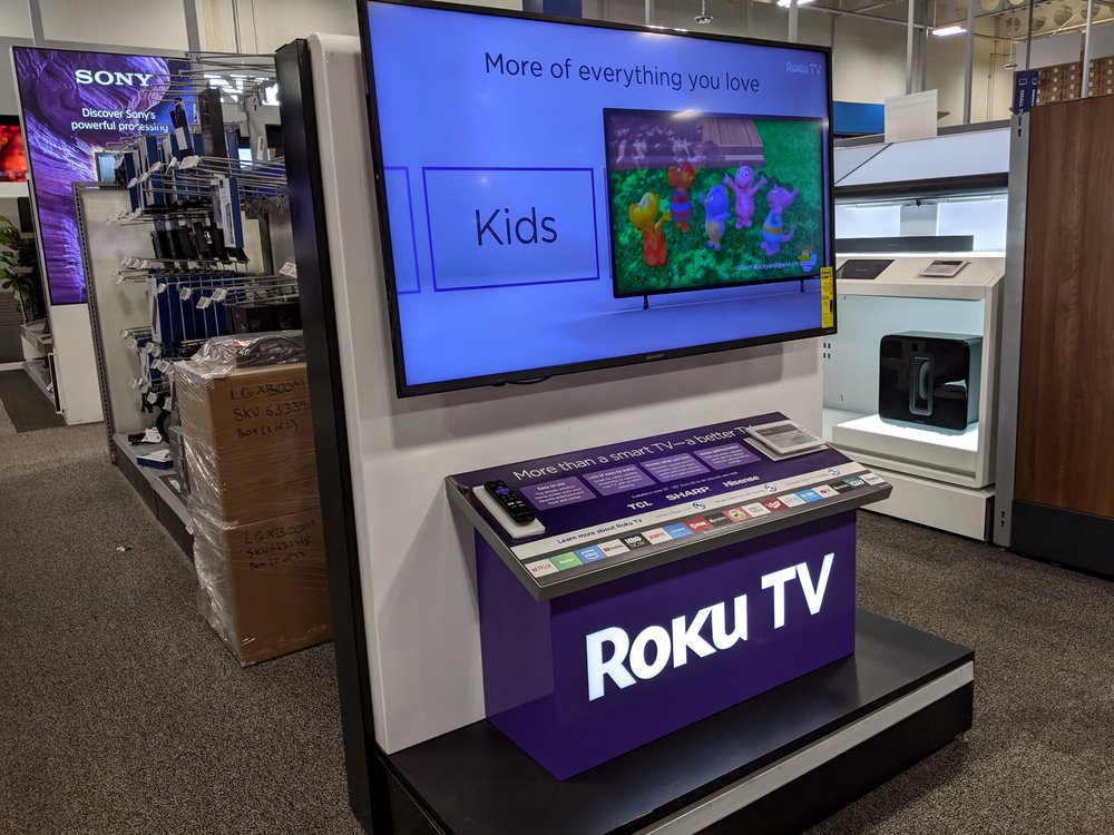 Izložba Roku TV-a u trgovini elektronikom prikazuje dječji TV program na ekranu, s promotivnim natpisima i daljinskim upravljačima na pultu ispod - prikazujući jedan od najboljih modela poput Hisense TV-a, popularnog zbog pozitivnih iskustava.