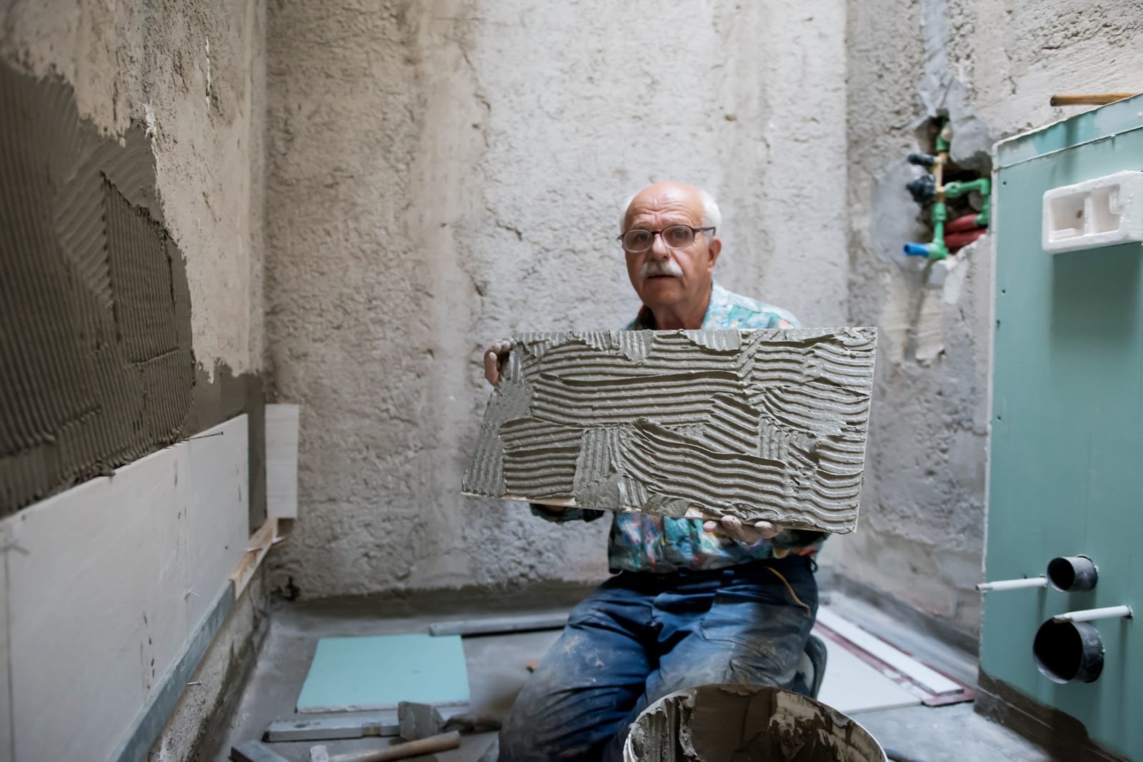 Stariji muškarac kleči na podu kupaonice koja se renovira, držeći veliku pločicu prekrivenu fleksibilnim ljepilom za pločice, okružen građevinskim materijalom.