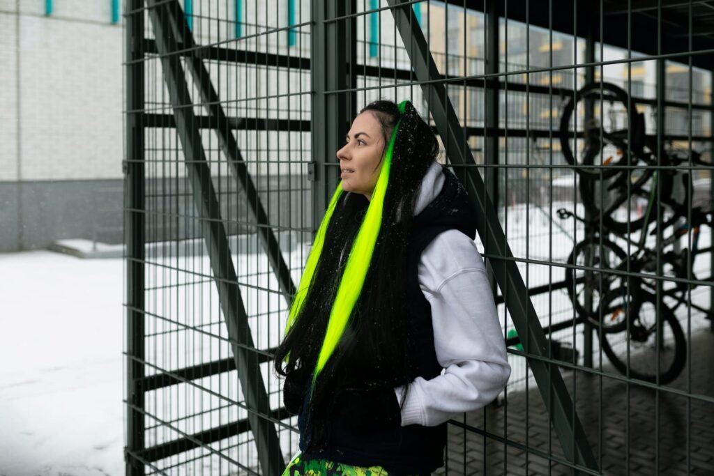 Žena s dugim crnim i neonsko žutim ekstenzijama za kosu stoji uz metalnu ogradu na snježnom vanjskom prostoru, pokazujući elegantan dizajn i prednje dijelove ograde. Bicikli su parkirani u pozadini.
