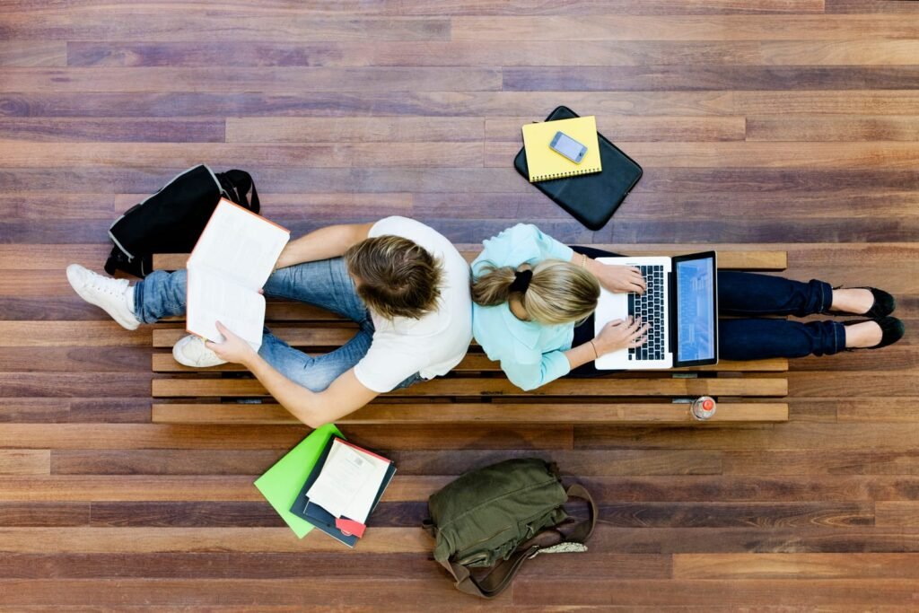 Dvoje ljudi sjedi na drvenoj klupi, jedan čita knjigu, a drugi koristi laptop; studijski materijali i torbe razbacane okolo sugeriraju da istražuju najbolje fakultete ili možda sastavljaju listu TOP 7 fakulteta u Zagrebu.