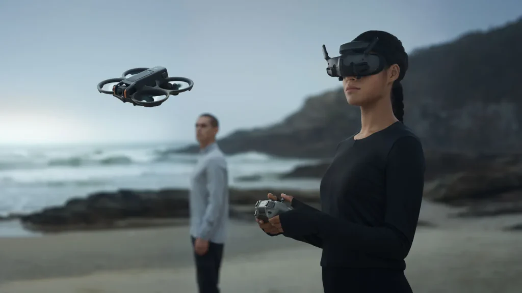 Žena s VR naočalama upravlja dronom s daljinskim upravljačem na plaži, dok muškarac stoji u pozadini blizu obale - idealna scena za sve koji traže preporuke za dronove ili odlučuju kupiti dron.