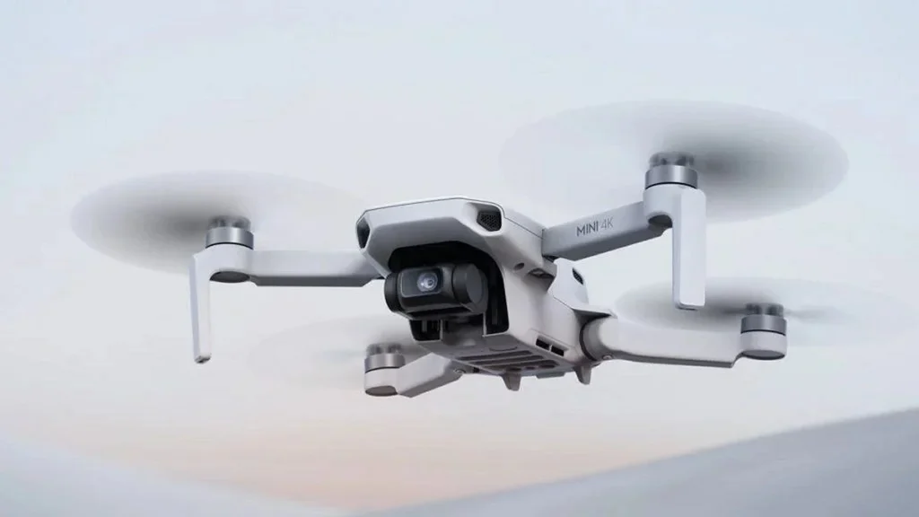Kompaktni bijeli kvadkopter dron s kamerom leti zrakom na neutralnoj, svijetloj pozadini - idealno za one koji traže preporuke ili pretražuju među najboljim dronovima.