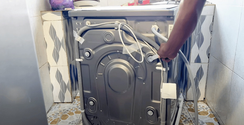 Osoba instalira ili popravlja Hisense perilicu rublja, rukuje njezinim crijevima i stražnjom pločom u popločanoj praonici rublja - idealno za sve koji traže iskustva ili recenzije o postavljanju ili održavanju.