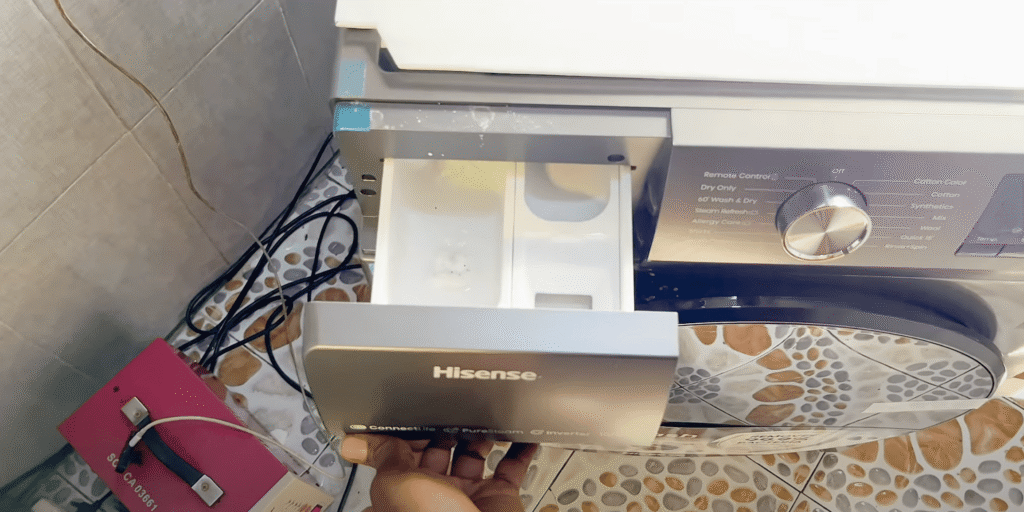 Ruka otvara ladicu za deterdžent Hisense perilice rublja u popločanoj prostoriji za pranje rublja, s vidljivim kabelima i ružičastom kutijom za napajanje u blizini - savršeno za one koji traže iskustvo ili recenziju ovog uređaja.