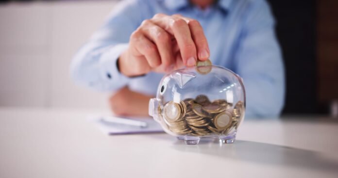 Osoba stavlja novčić u prozirnu kasicu punu novčića, sjedeći na bijelom stolu s notesom i olovkom u pozadini - savršeno za planiranje štednje ili korištenje najboljeg kalkulatora za praćenje financija.