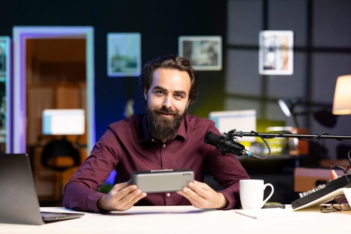 Muškarac s bradom sjedi za stolom s prijenosnim računalom, mikrofonom i šalicom za kavu, držeći nešto što izgleda kao Bluetooth zvučnik, u modernom uredskom ili studijskom okruženju.