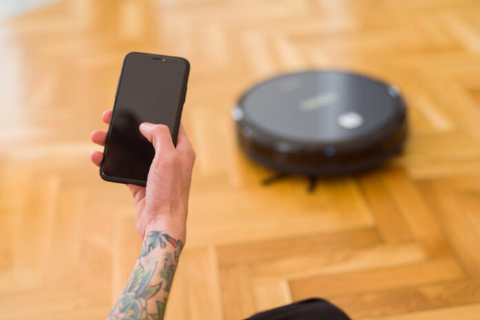 Osoba koja drži pametni telefon upravlja robotskim usisavačem na drvenom podu, bez napora održavajući čistoću doma.