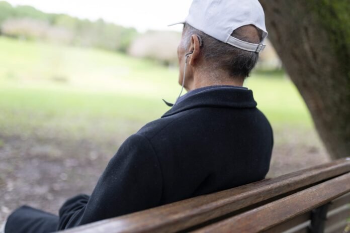 najbolji slusni aparat Stariji muškarac u bijeloj kapi i tamnom kaputu sjedi na drvenoj klupi vani, okrenut prema travnatim drveću i slušajući uz pomoć Slušnih aparata, uživajući u čistom zvuku - preporuka za sve koji traže najbolje slušalne aparate.