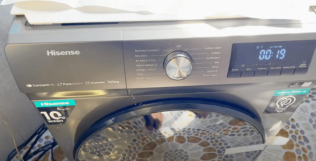 Hisense perilica rublja s prednjim punjenjem, digitalnim zaslonom, upravljačkom tipkom i označenim tipkama stoji na popločanom podu. Zaslon prikazuje preostalo vrijeme od 00:19 - idealno za one koji traže prava iskustva i recenzije prije kupnje.