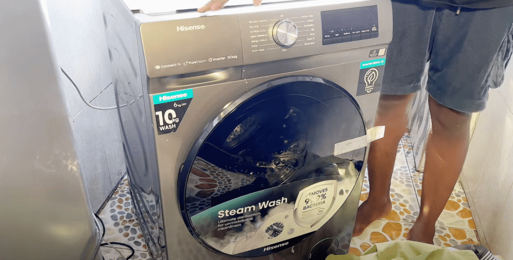 Osoba upravlja Hisense perilicom rublja s prednjim punjenjem na paru na popločanom podu, s vidljivom parom unutar bubnja, što pokazuje pravo iskustvo s ovim modernim uređajem.