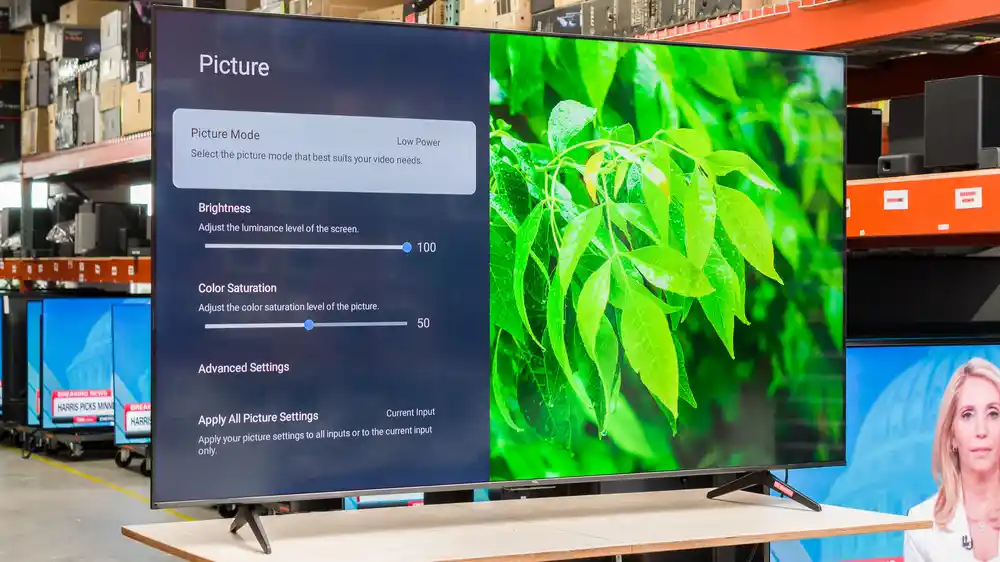 Veliki televizor ravnog ekrana na stalku prikazuje izbornik postavki s lijeve strane i krupni plan zelenog lišća s desne strane, s policama i monitorima u pozadini - savršeno za svakoga tko razmišlja o kupnji ili piše recenziju TCL televizora.