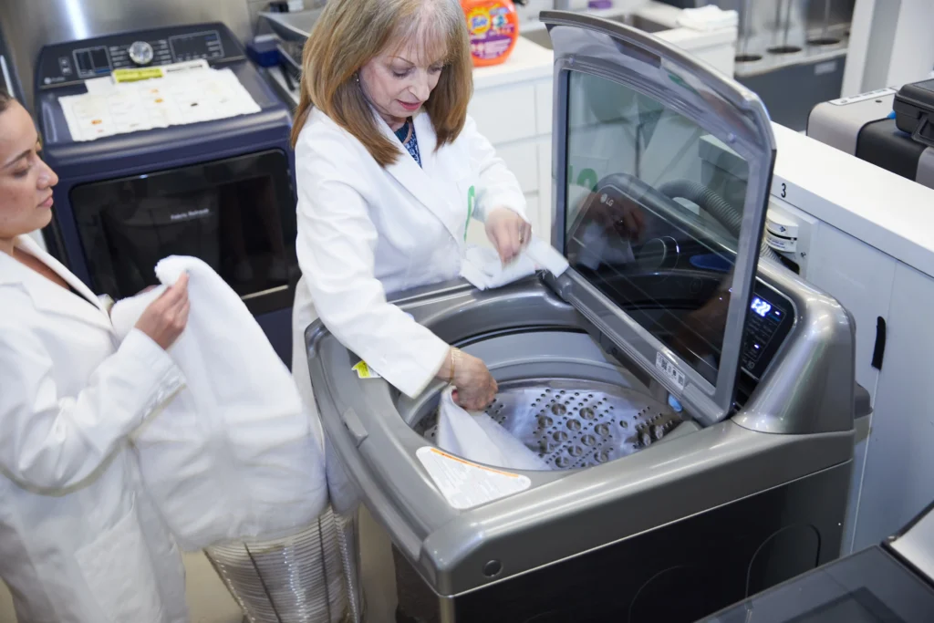 Dvije žene u laboratorijskim kutama stavljaju bijele ručnike u rublje s gornjim punjenjem u laboratorijskom okruženju, nakon svake preporuke za točno testiranje.