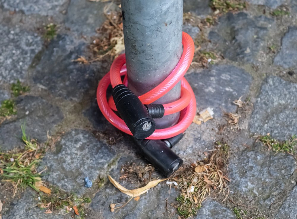 Crveni Lokot za bicikl je omotan oko metalnog stupa, ali ne osigurava bicikl. Brava se nalazi na kamenom pločniku s travom u blizini, što jasno daje do znanja zašto je pronalazak najboljeg lokota uvijek preporuka za bicikliste.